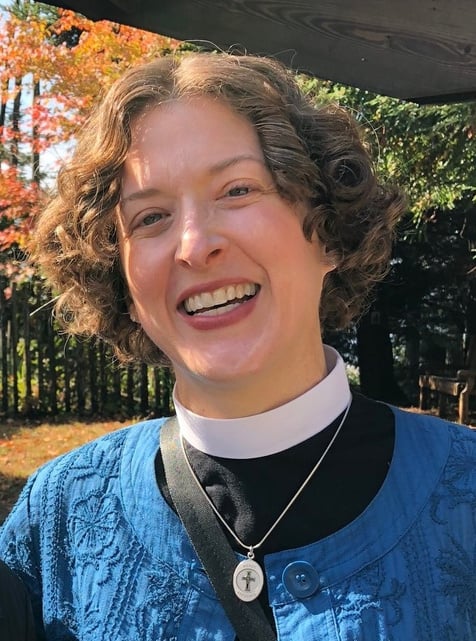 A pale-skinned woman wearing a cross necklace and a shirt with clergy collar is smiling for the camera