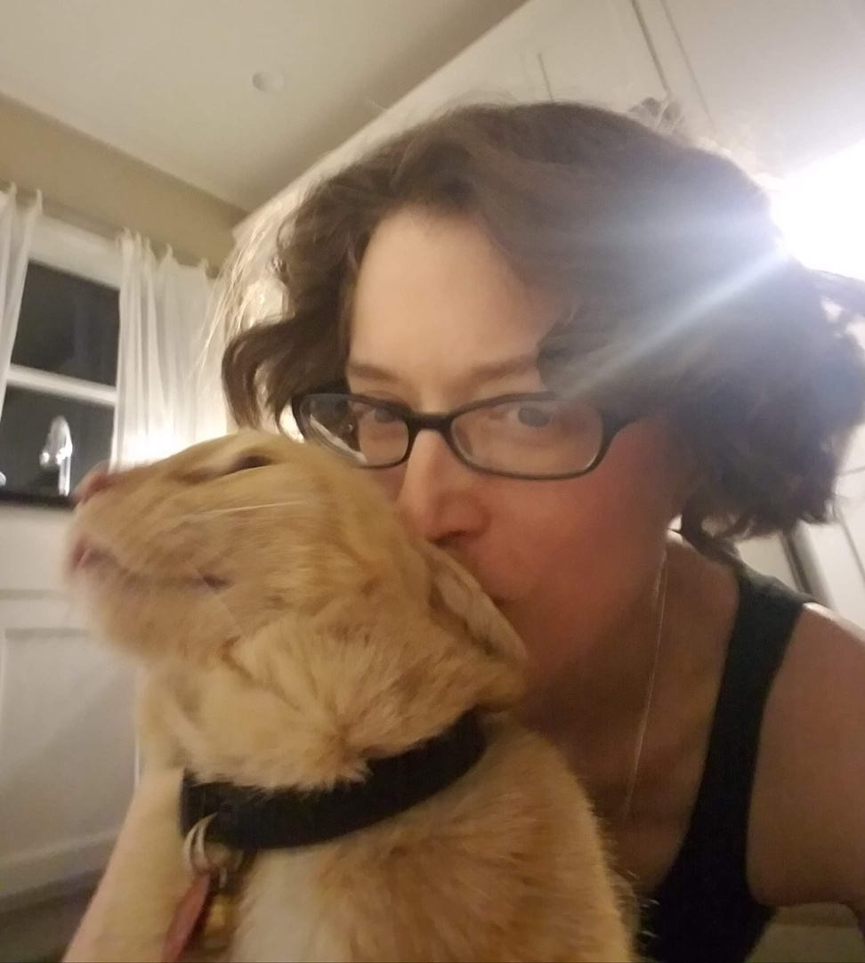 A woman wearing glasses kisses a happy orange tabby cat on the top of the head