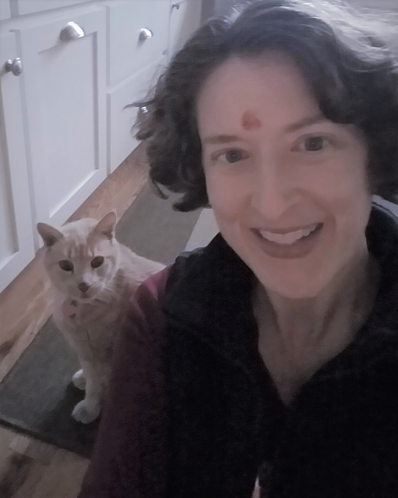 A woman kneeling on the floor, smiling, next to an orange tabby cat who is looking into the camera.